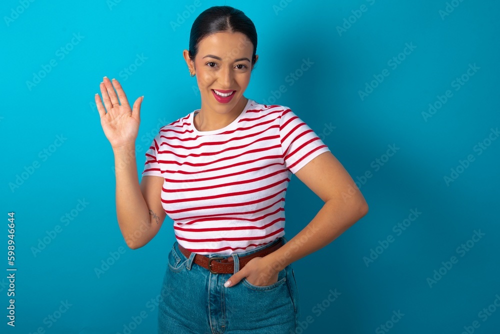 beautiful woman wearing striped T-shirt over blue studio background waiving saying hello or goodbye happy and smiling, friendly welcome gesture. beautiful woman wearing striped T-shirt over blue studio background waiving saying hello or goodbye happy and smiling, friendly welcome gesture.
