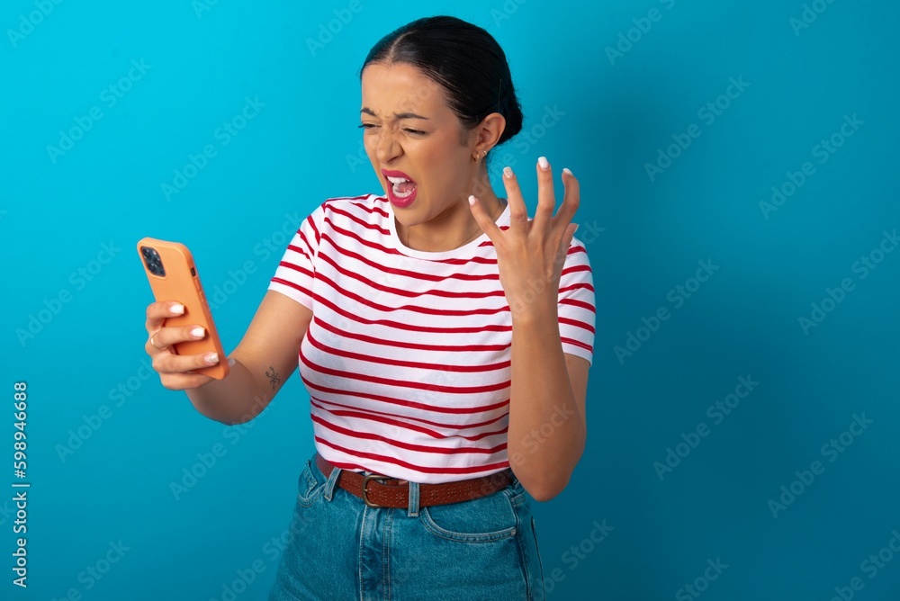 Photo of outraged annoyed beautiful woman wearing striped T-shirt over blue studio background holds cell phone, makes call, argues with colleague, expresses negative emotions. People and anger. Photo of outraged annoyed beautiful woman wearing striped T-shirt over blue studio background holds cell phone, makes call, argues with colleague, expresses negative emotions. People and anger.