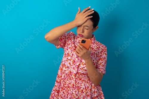 Upset depressed beautiful woman wearing floral dress over blue studio background makes face palm as forgot about something important holds mobile phone expresses sorrow and regret blames