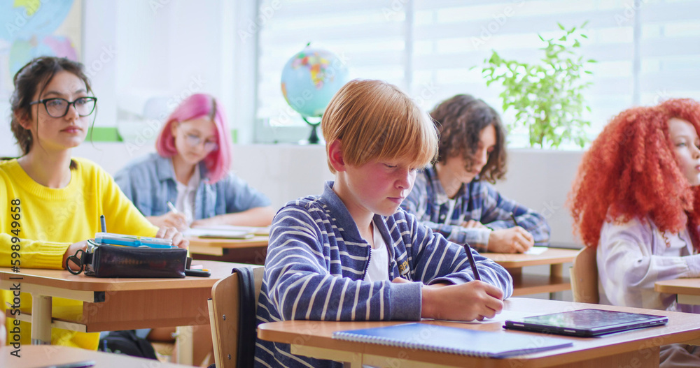 Portrait of nice small teen clever schoolboy sitting at desk in classroom and writing. Among kids. Inside class. Little boy learning in school and answering. Portrait of nice small teen clever schoolboy sitting at desk in classroom and writing. Among kids. Inside class. Little boy learning in school and answering.