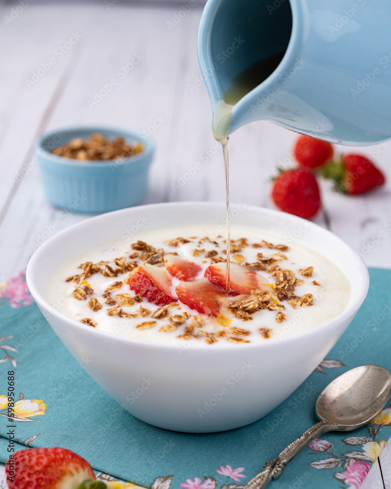 Yogurt with strawberry, granola and pouring honey in a bowl over white wooden table Yogurt with strawberry, granola and pouring honey in a bowl over white wooden table
