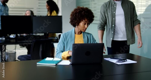Young businesswoman talking with a male coworkers while writing notes and using a laptop at her desk in an office