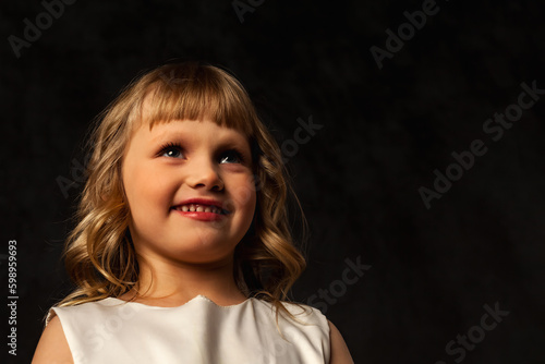 Face of cover baby doll girl 4-5 year old isolated at black background, smiling looking away. Adorable baby posing in shadow, studio shot. Theatre show performance concept. Copy ad poster text space