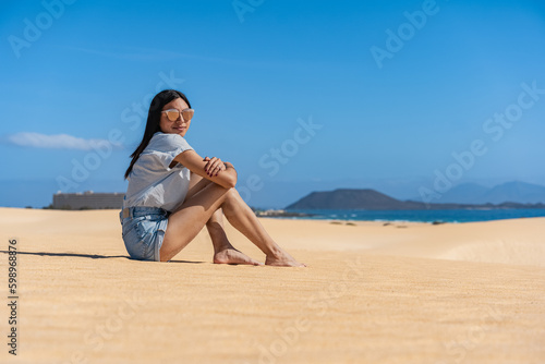 mujer morena de cabello negro sentada en la arena amarilla con ropa de color azul y gafas 