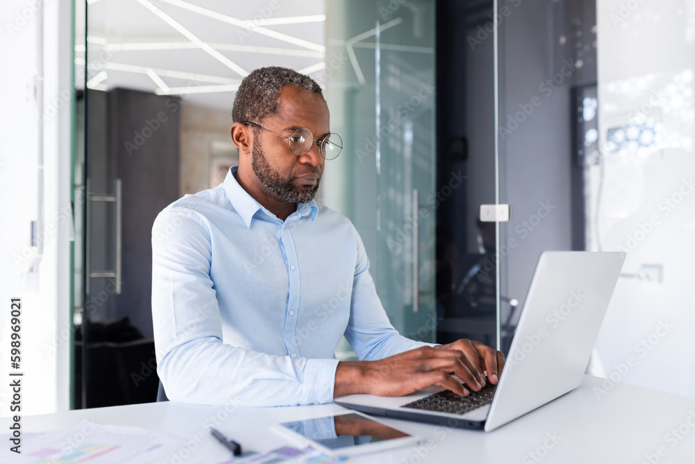 Serious and focused african american businessman in shirt working inside office with laptop, mature investor financier at workplace with documents graphs and charts. Serious and focused african american businessman in shirt working inside office with laptop, mature investor financier at workplace with documents graphs and charts.