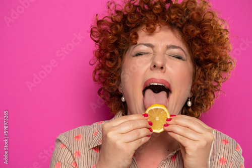 Portrait of curly red hair woman licking a slice of lemon.