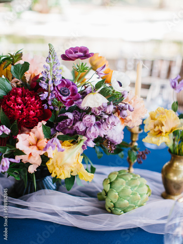 Bright decor of exotic flowers. Bouquet with artichoke, delphinium, iris, lily of the tongue, tulips. Blue tablecloth, draped with white tulle.