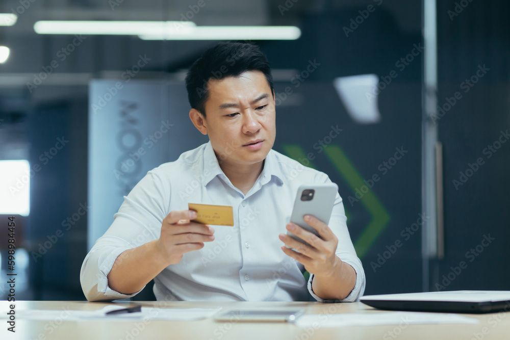 Φωτογραφία Worried young asian man sitting at desk in office and holding phone and blocked credit card, financial problems, unsuccessful shopping, money transfer