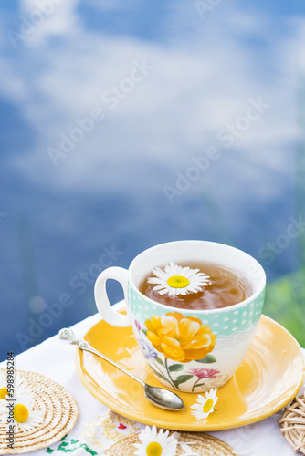 Herbal tea in teacup decorated with flowers outdoors in background of blue sky with clouds, healthy herbal drink with honey, tea drinking concept