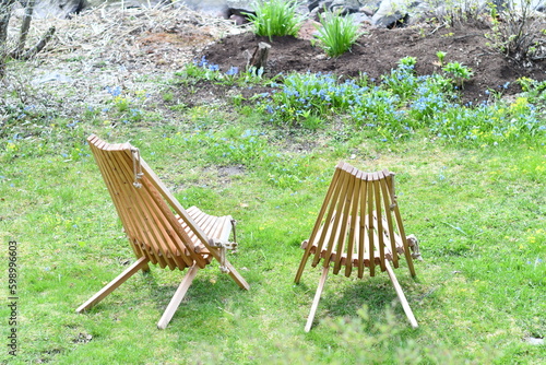 Two wooden Garden chairs on the grass. Small green garden and a flower bed with blue flowers and greenery on the background. Wooden benches facing away.