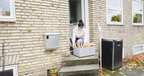 Woman in a protective face mask bringing a grocery delivery box into her home from her doorstep