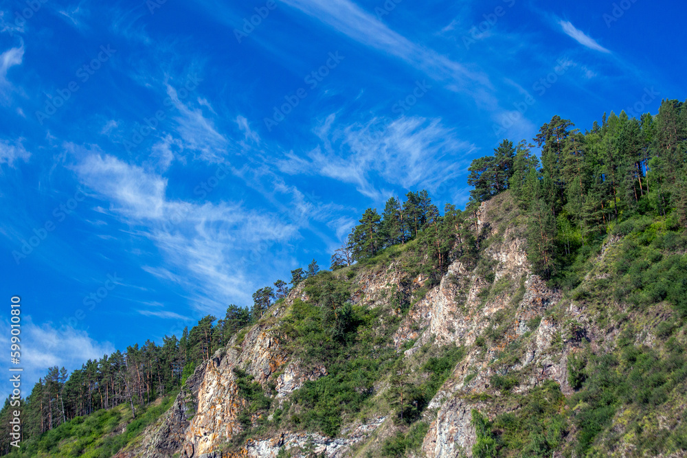 Picturesque rocks covered with coniferous forest against a blue sky with white clouds.