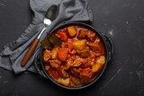 Beef meat stew with potatoes, carrot and delicious gravy in black casserole pot with bay leaves with spoon on black dark rustic concrete background from above . Generative AI