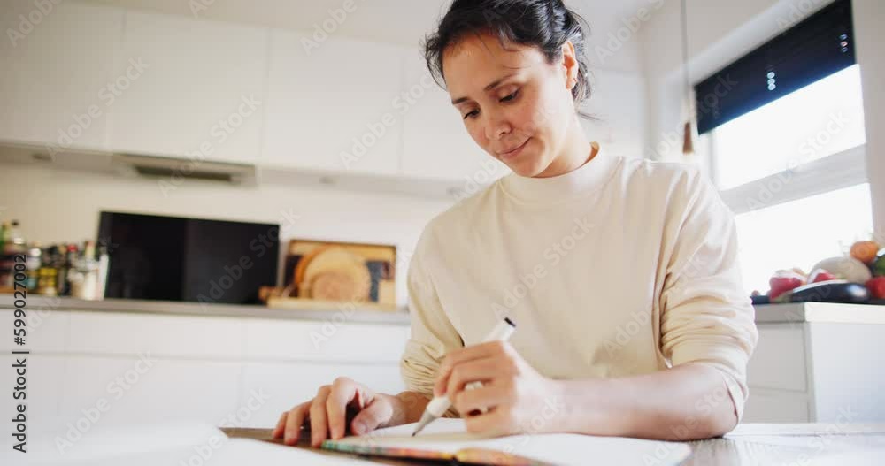 Smiling young woman sketching designs in a book while sitting at a table in her kitchen at home Smiling young woman sketching designs in a book while sitting at a table in her kitchen at home