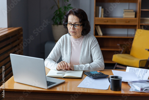 Middle aged senior woman sit with laptop and paper document. Pensive older mature lady reading paper bill pay online at home managing bank finances calculating taxes planning loan debt pension payment