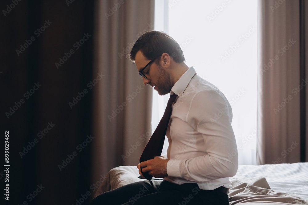 Young businessman taking off his tie while sitting on a bed in a hotel room. Young businessman taking off his tie while sitting on a bed in a hotel room.