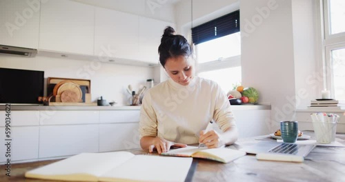 Young woman smiling while sitting at a table in her kitchen at home and sketching designs in a book