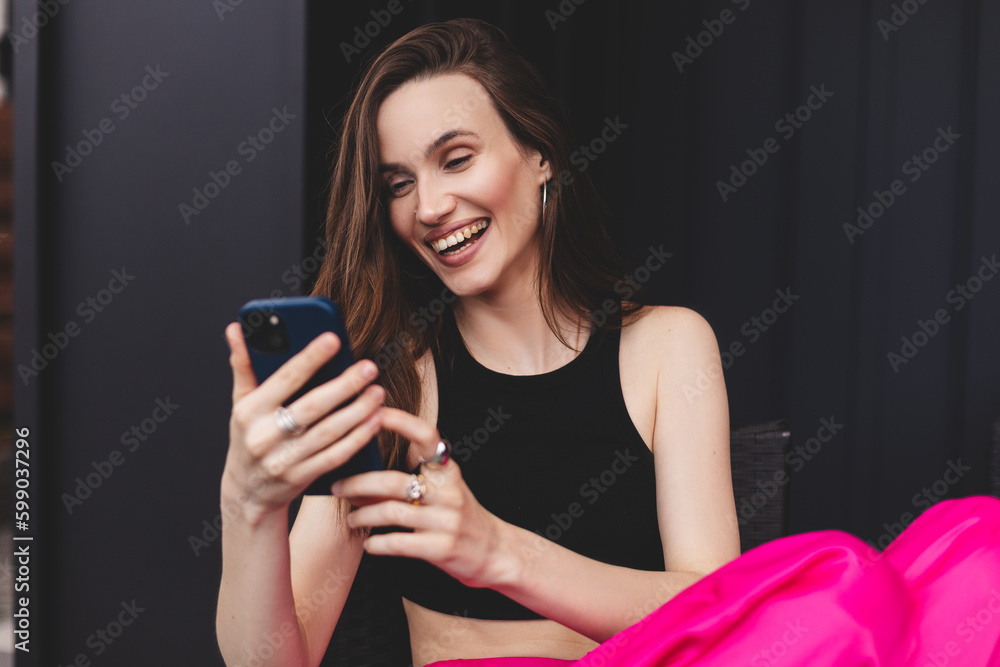 Delighted brunette girl show wow emotions and using mobile phone sitting on the street at home outdoors on black wall. Surprised girl surfing internet on mobile phone, wear pink pants and black top. Delighted brunette girl show wow emotions and using mobile phone sitting on the street at home outdoors on black wall. Surprised girl surfing internet on mobile phone, wear pink pants and black top.