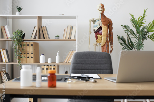 Obraz na płótnie Focus on human anatomy model on stand near office shelving with books and pills bottles at consulting room with writing desk on blurred foreground