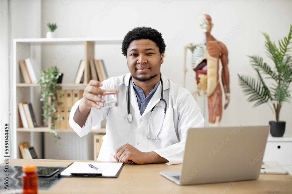 Positive multicultural physician in lab coat showing glass of water while sitting at writing desk with laptop and documents in doctor's office. General practitioner recommending staying hydrated. Positive multicultural physician in lab coat showing glass of water while sitting at writing desk with laptop and documents in doctor's office. General practitioner recommending staying hydrated.