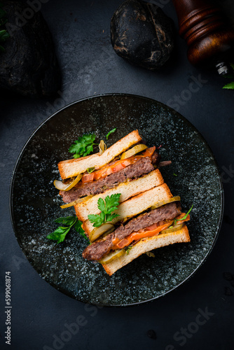 Sandwich with beef cutlet, pickled cucumber, tomatoes, lettuce and greens on dark background.