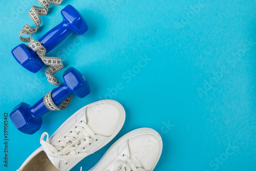The concept of fitness, sports, weight loss. Top view photo of white sports shoes, measuring tape and blue dumbbells on isolated pastel blue background with empty space.