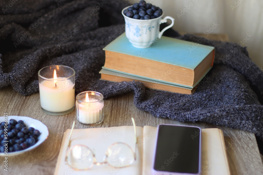Cup of blueberries, set of books, phone, eyeglasses, lit candles and blanket on the table. Hygge at home. Selective focus. Cup of blueberries, set of books, phone, eyeglasses, lit candles and blanket on the table. Hygge at home. Selective focus.
