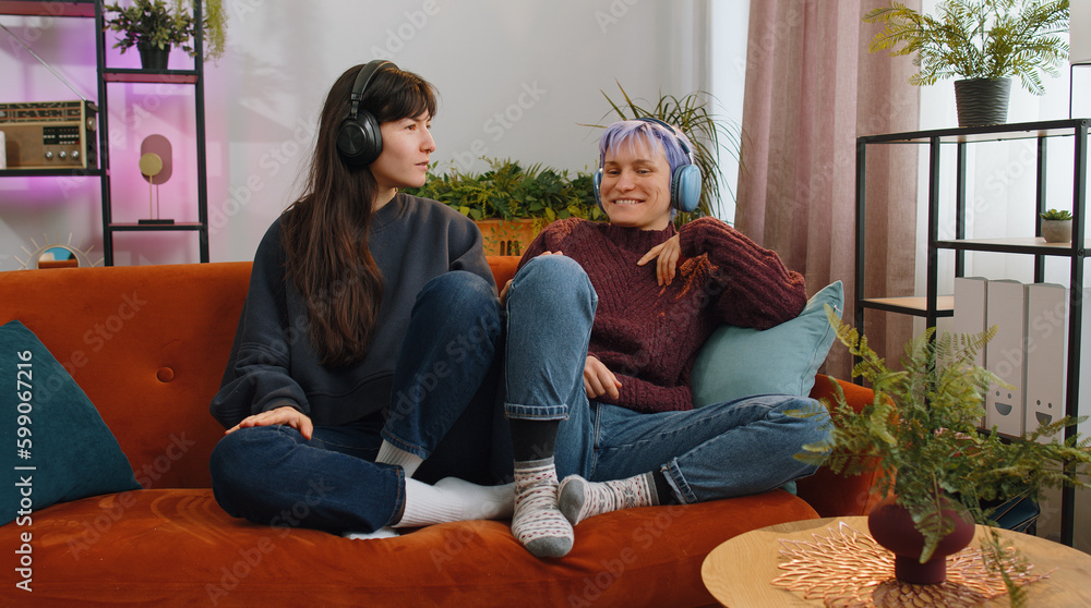 Happy relaxed overjoyed young lesbian women family couple or girls friends in wireless headphones dancing choosing listening favorite energetic disco rock n roll music song on home couch. LGBT people Happy relaxed overjoyed young lesbian women family couple or girls friends in wireless headphones dancing choosing listening favorite energetic disco rock n roll music song on home couch. LGBT people