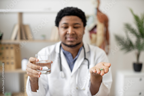 Focus on green round pill being held by multiracial male doctor sitting at writing desk with laptop on office background. Professional medical worker giving medicine and glass of water for treatment.