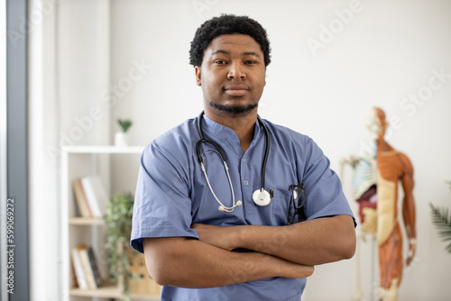Handsome multiracial male specialist wearing blue scrubs and stethoscope posing with arms crossed in doctor's office interior. Medical practitioner taking break from patients examination in clinic.