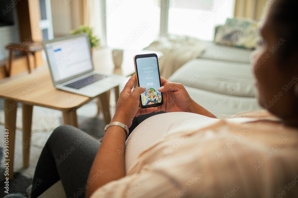 Young pregnant latina woman using a diet app on her smart phone while sitting on a couch in the living room of her home Young pregnant latina woman using a diet app on her smart phone while sitting on a couch in the living room of her home