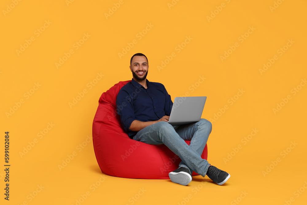 Smiling young man working with laptop on beanbag chair against yellow background Smiling young man working with laptop on beanbag chair against yellow background