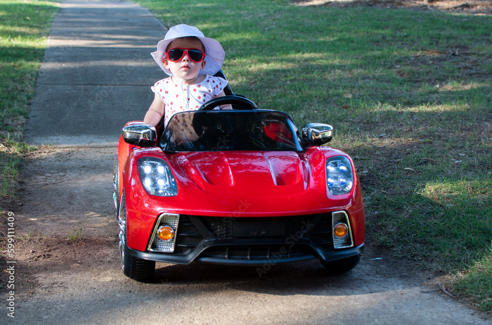 Fototapeta premium Caucasian baby in a white hat riding in an electric convertible red sport car. child driving a car on a summer day