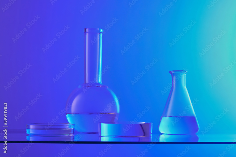 Front view of lab glassware containing colorless liquid and transparent podium displayed on blue background. Pedestal for cosmetic product presentation with laboratory concept. Front view of lab glassware containing colorless liquid and transparent podium displayed on blue background. Pedestal for cosmetic product presentation with laboratory concept.