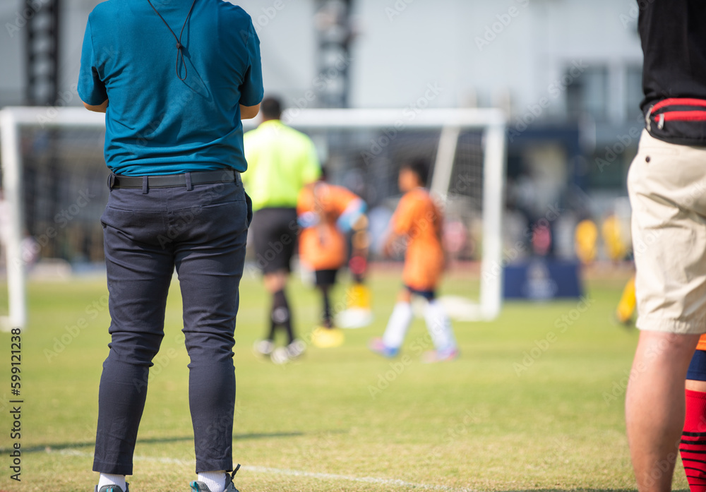 Fototapeta premium Father standing and watching his son playing football in a school tournament on a clear sky and sunny day. Sport, outdoor active, lifestyle, happy family and soccer mom and soccer dad concept.