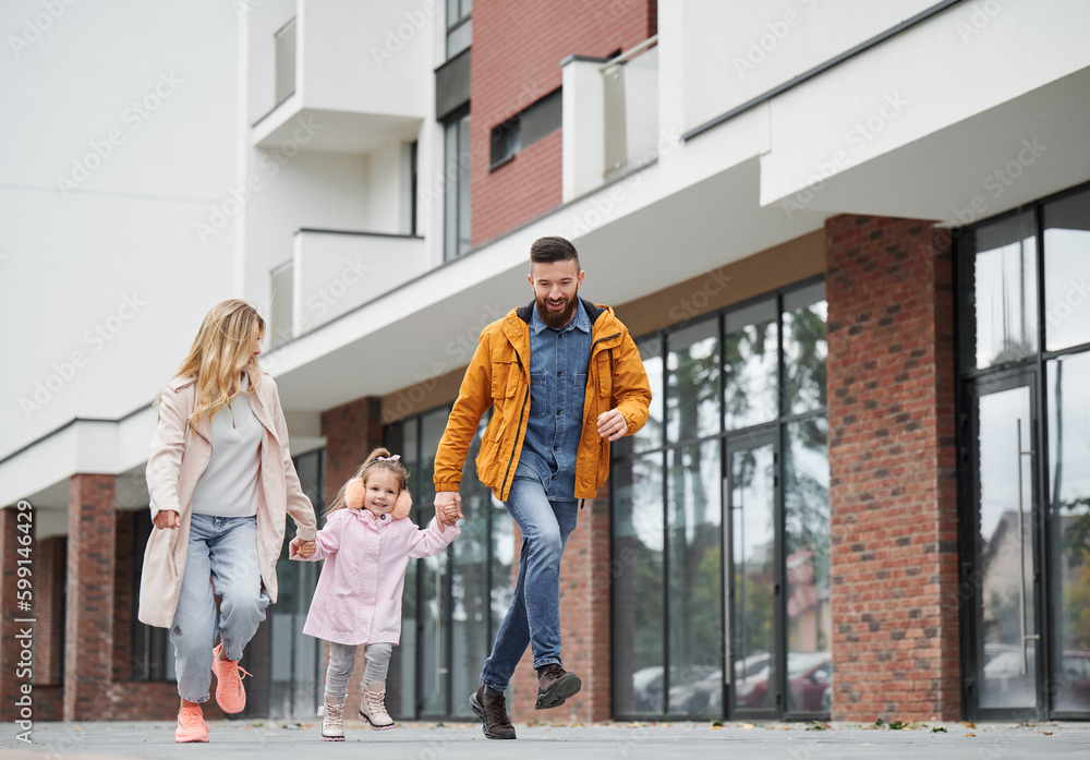 Full length of man and woman holding hands of little girl while walking down the street in new urban district. Happy loving family with child enjoying stroll outdoors near residential building. - obrazy, fototapety, plakaty 