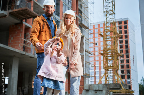 Daughter holding apartment keys and smiling while standing next to parents outside building under construction. Happy family homeowners posing on the street at construction site.