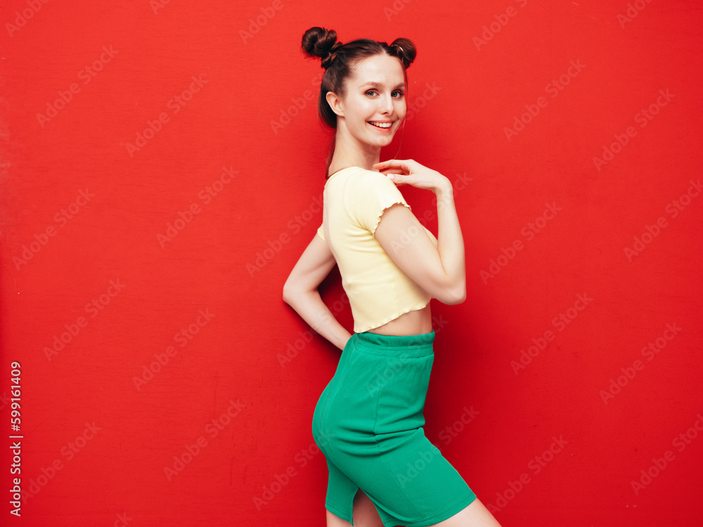 Young beautiful smiling female in trendy summer yellow t-shirt and green skirt. Carefree woman with two horns hairstyle posing near red wall in studio. Positive model having fun. Cheerful and happy Young beautiful smiling female in trendy summer yellow t-shirt and green skirt. Carefree woman with two horns hairstyle posing near red wall in studio. Positive model having fun. Cheerful and happy