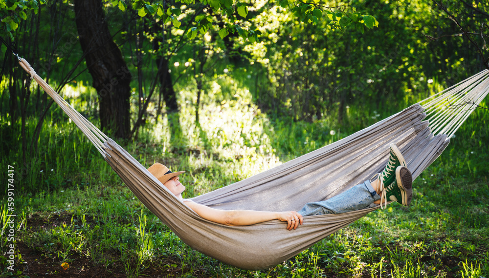 Young happy caucasian woman in a hat lying in a hammock in a green garden enjoying a summer holidays