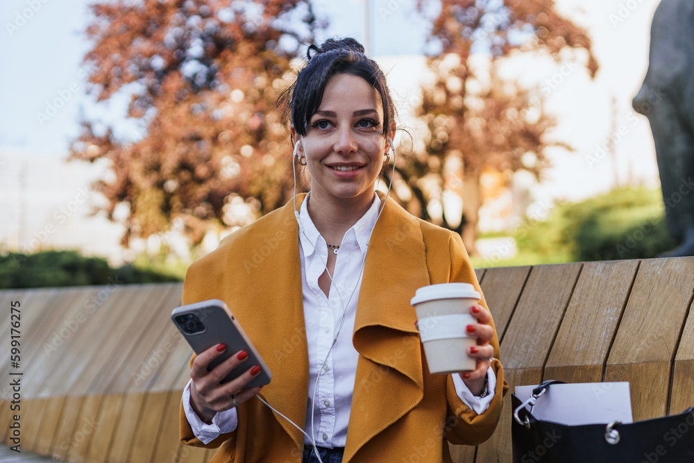 Fototapeta premium Portrait of a business woman on a coffee break listening to a podcast in a park,drinking coffee. Woman sitting on a bench in a park looking away from camera, it's fall and she's wearing a coat