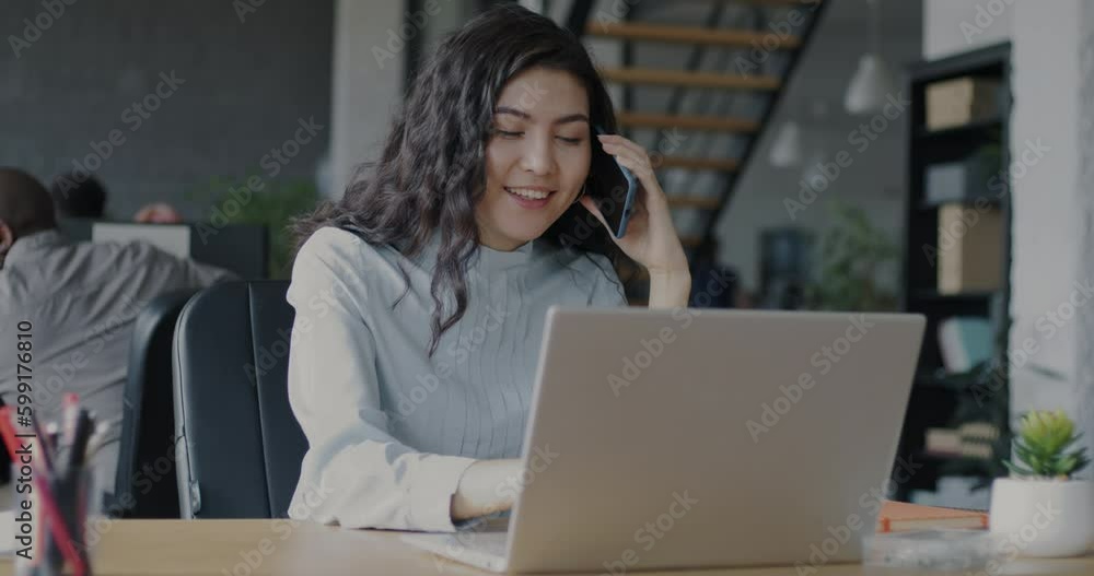 Cheerful young woman manager speaking on mobile phone and working with laptop in coworking office. Business communication and technology concept. Cheerful young woman manager speaking on mobile phone and working with laptop in coworking office. Business communication and technology concept.