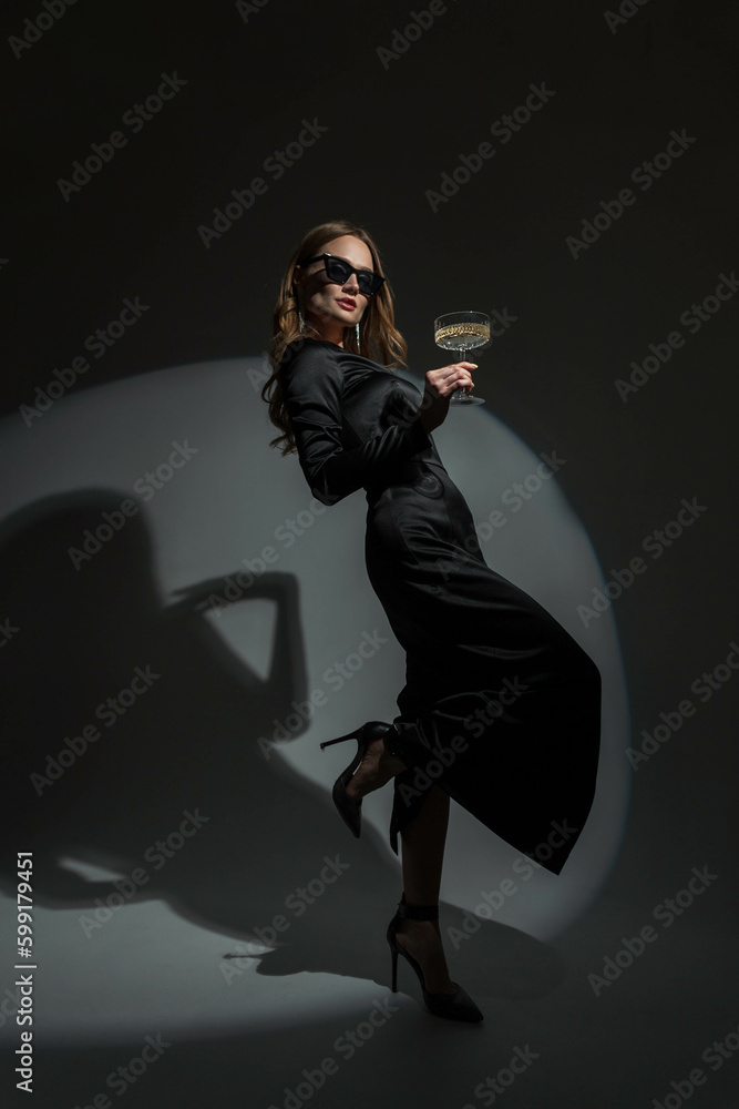 Obraz premium Gorgeous beautiful stylish woman with cool glasses in a trendy black evening dress with heels holds a glass of champagne and celebrates the event on a dark background. Pretty chic Lady at the party