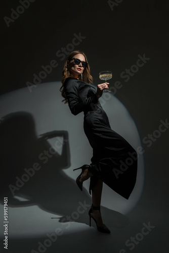 Foto Gorgeous beautiful stylish woman with cool glasses in a trendy black evening dress with heels holds a glass of champagne and celebrates the event on a dark background