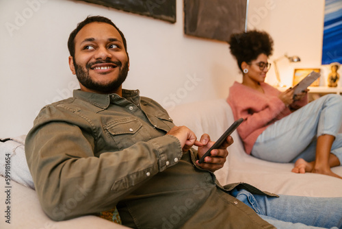 Young indian man using mobile phone while sitting on couch with african woman who's reading book © Drobot Dean
