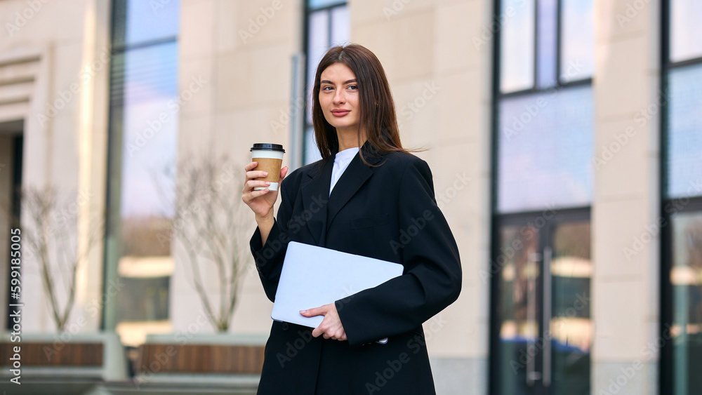 A dedicated professional in a stylish business suite, holding a coffee cup and a laptop as she stands against a backdrop of city skyscrapers A dedicated professional in a stylish business suite, holding a coffee cup and a laptop as she stands against a backdrop of city skyscrapers