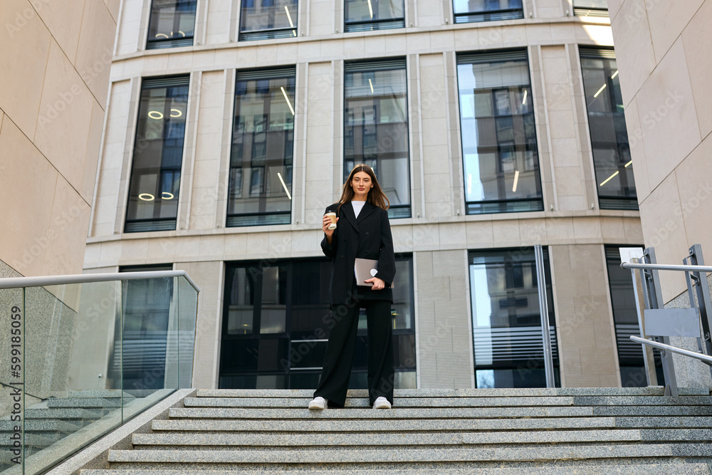 A stylish business woman in a black suit is standing on the street against the background of a business center and drinking coffee A stylish business woman in a black suit is standing on the street against the background of a business center and drinking coffee