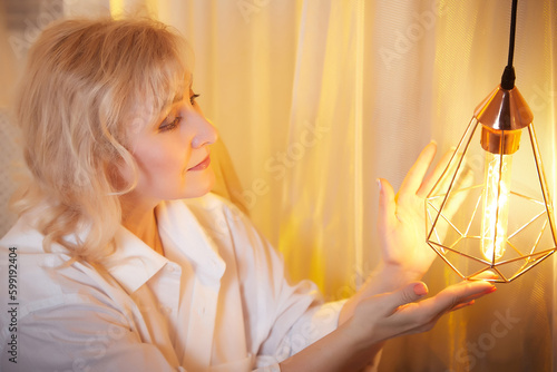 Adult mature woman of 40-60 years old with lapm in casual dress of white shirt in calm cozy evening atmosphere room. Interior with transparent curtains and soft light of warm lamps in the loft style photo