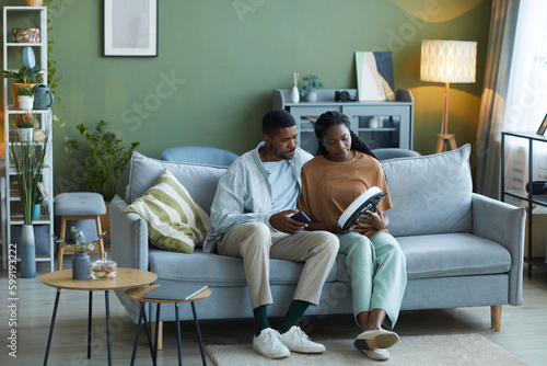 African American couple setting the robot vacuum cleaner before housework while sitting on sofa in the living room
