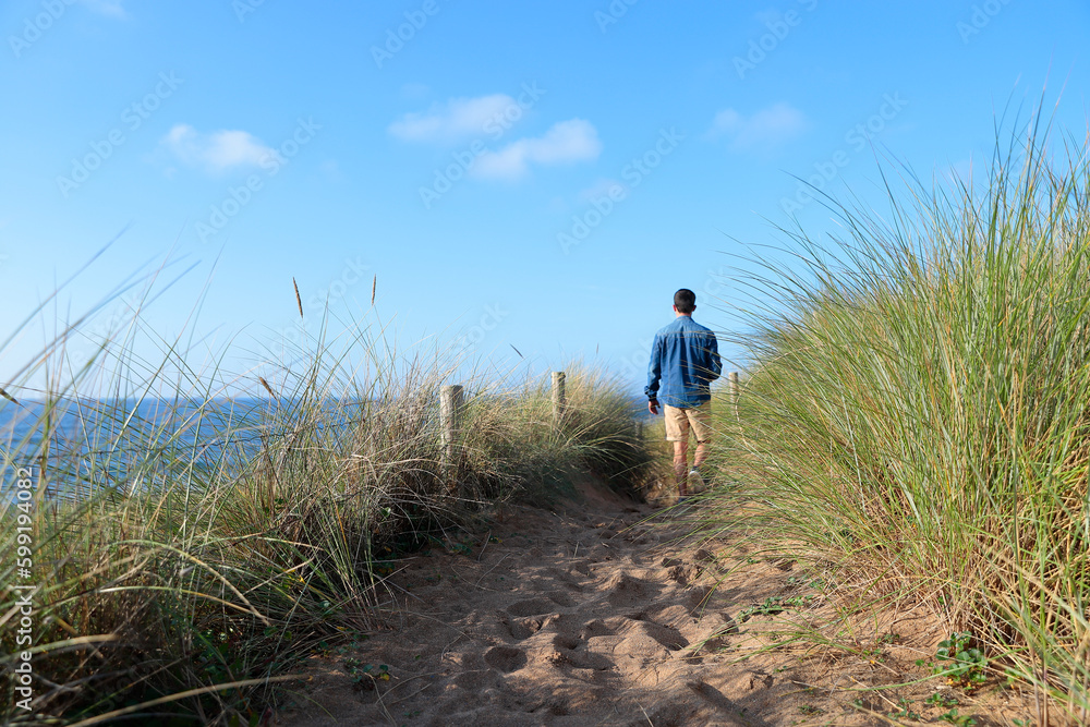 Coastal path with plants on the sides and illuminated by lateral sunlight, an unfocused person walks along it in the background, concept of lifestyle and tranquility.