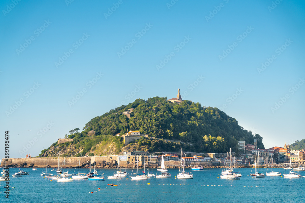 Fototapeta premium SAN SEBASTIAN, Spain July 08 2022: View of Santa Clara Island. Boats docked in the middle of La Concha Bay. Beautiful travel destination in north of Spain. Jesus Christ Statue on top of the hill.